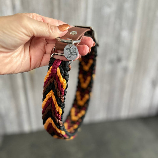 Ein Hundehalsband aus Paracord wird in einer Hand gehalten. Die Farben sind weinrot, orange, braun und schwarz